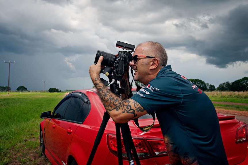 A man wearing a t-shirt looks through camera. Behind him is a red care, grass and a storm cloud. 