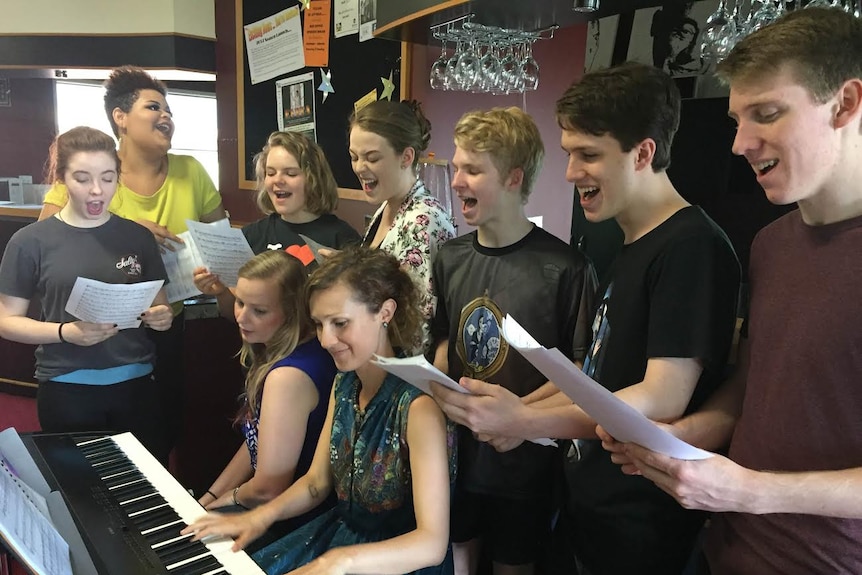 A groups of teenage students sing around a piano