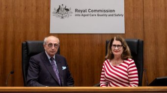 A man in a suit and a woman in a red and white stripped shirt sit behind a desk in court and pose for a photo.