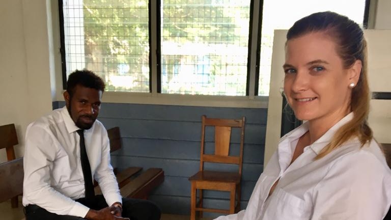 Lady sitting in foreground, with local Solomon Islander man sitting in background , inside building.