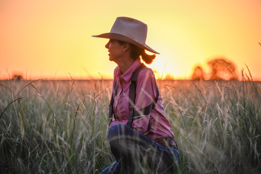 portrait of a woman wearing a widebrim hate and check shirt in a grass field as the sun sets behind her