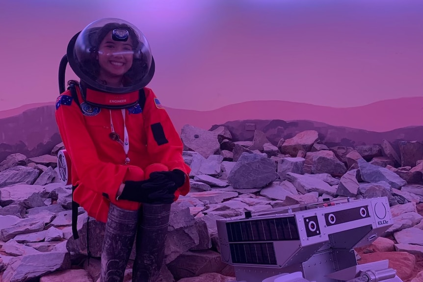 A woman in orange astronaut gear sits on rocky terrain next to rover
