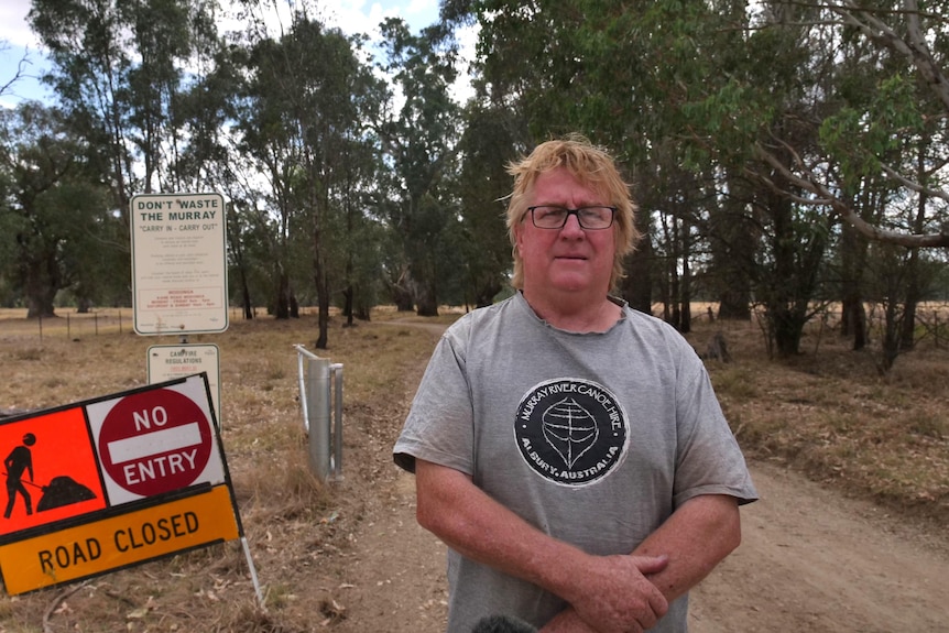A man stands on a dirt road with a road closed sign behind him 