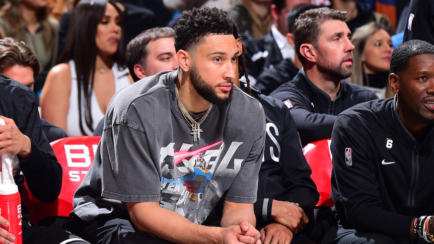 Australian NBA basketballer Ben Simmons sits on the sidelines during a game, wearing a T-shirt rather than a team uniform. 