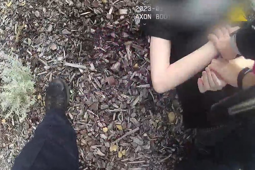 A close-up shot of a teenage boy in a black t-shirt lying on the ground with his arms held behind his back by a police officer.