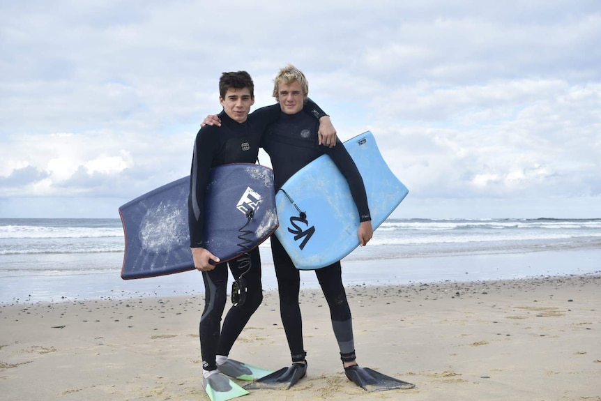 Two teenage boys standing in front of beach with surfboards