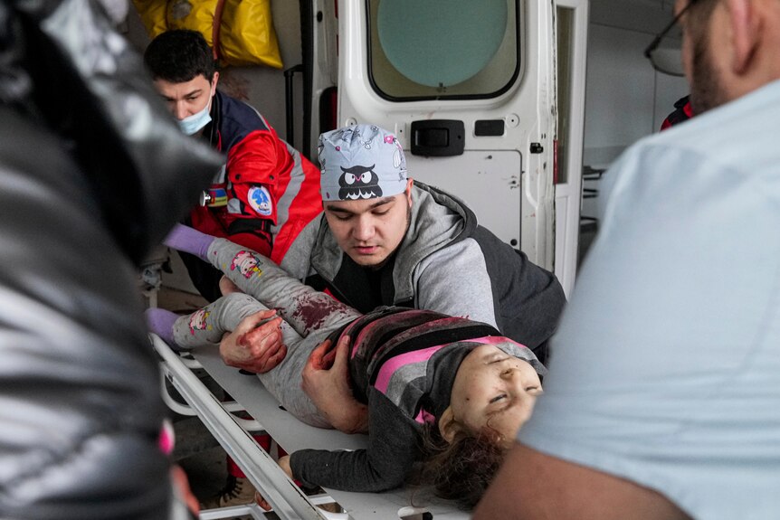 Medics moving a young girl out of a van. She has blood on her legs and her eyes are closed.