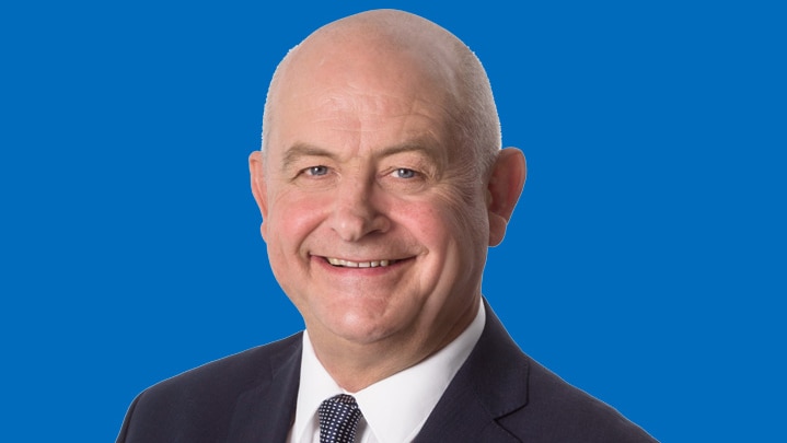 Jeremy Hearn smiles into the camera against a blue background. He wears a dark suit and tie, with a white shirt.