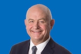 Jeremy Hearn smiles into the camera against a blue background. He wears a dark suit and tie, with a white shirt.