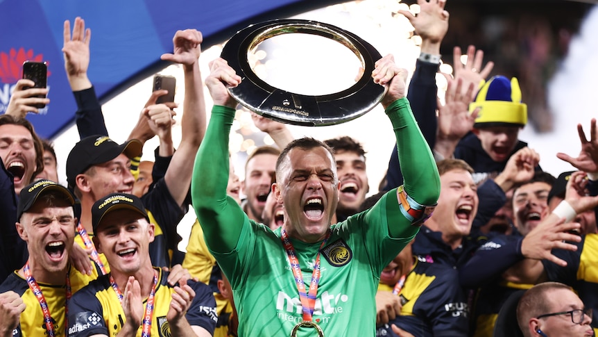 A Central Coast Mariners A-League Men player holds up the championship trophy with his teammates behind him.
