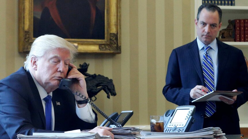 Donald Trump sits at desk talking on phone, Reince Priebus stands in the background. January 28, 2017.