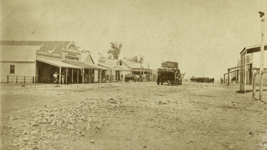 Bullocks pull a loaded cart down the main street, lined both sides with shops and buildings, in black and white.
