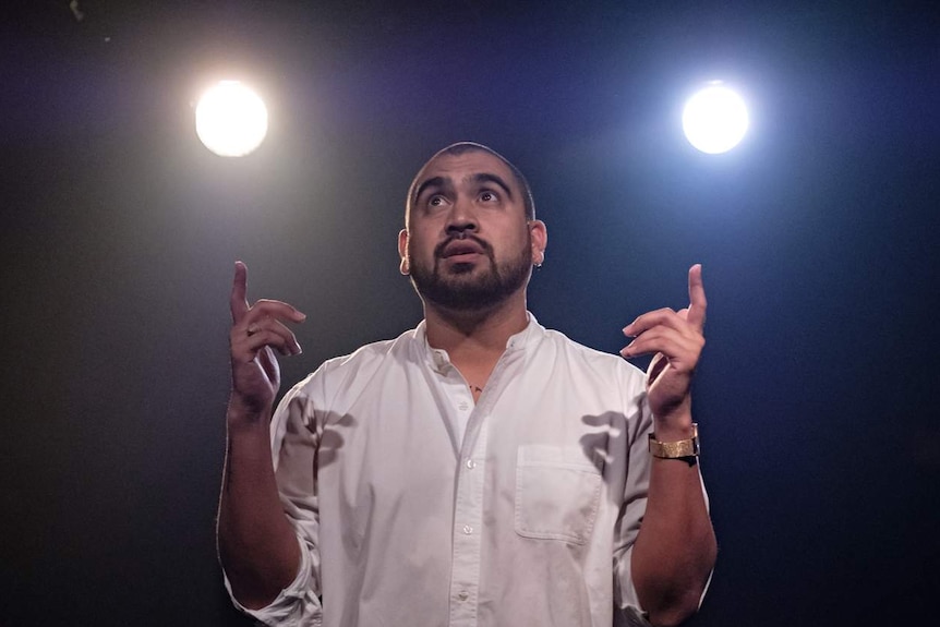 Man in his 20s with close-shaved head and wearing white shirt on stage backlit by two spots.