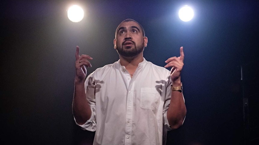 Man in his 20s with close-shaved head and wearing white shirt on stage backlit by two spots.