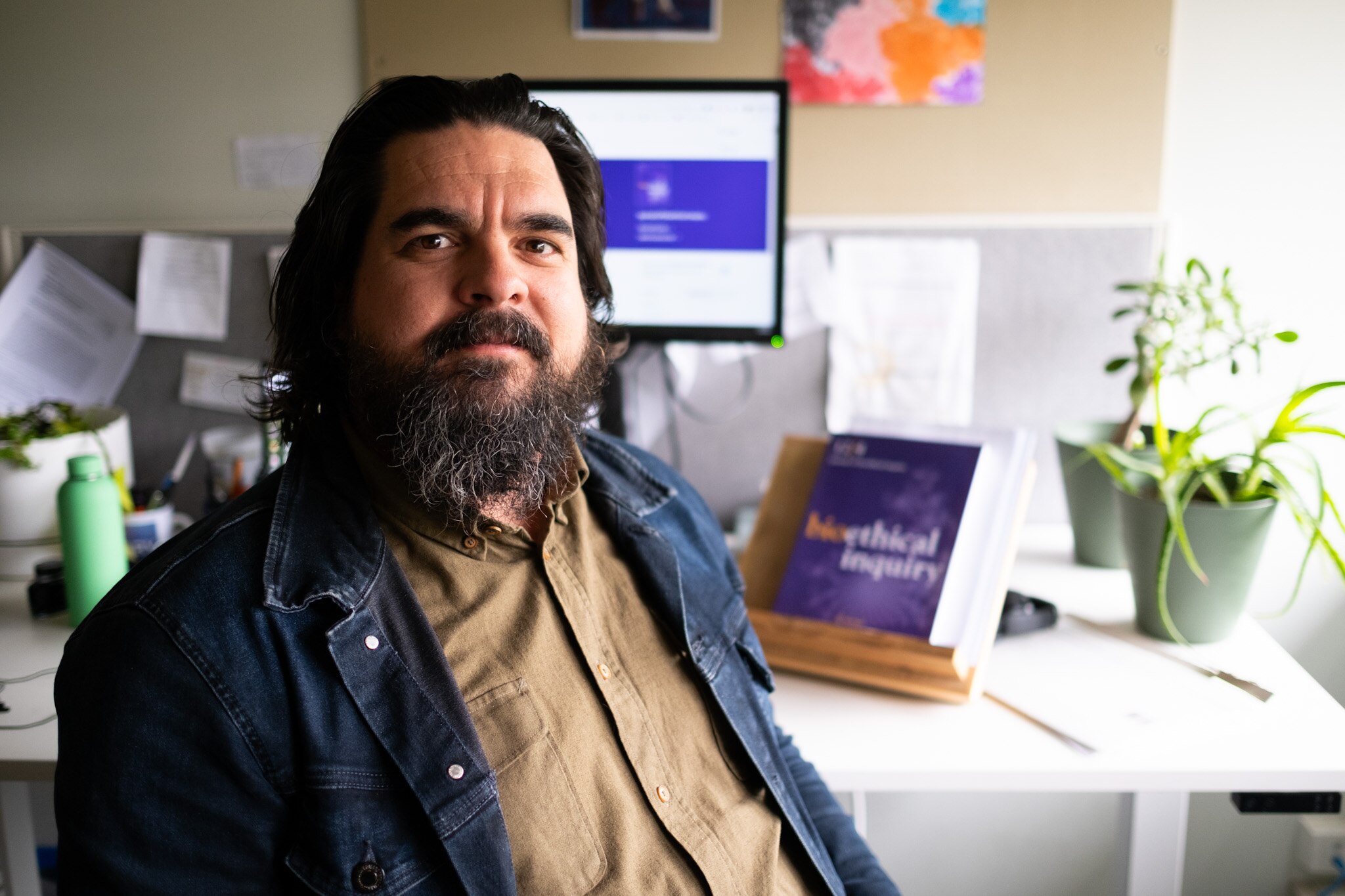 A man sits facing the camera with a computer and desk behind him