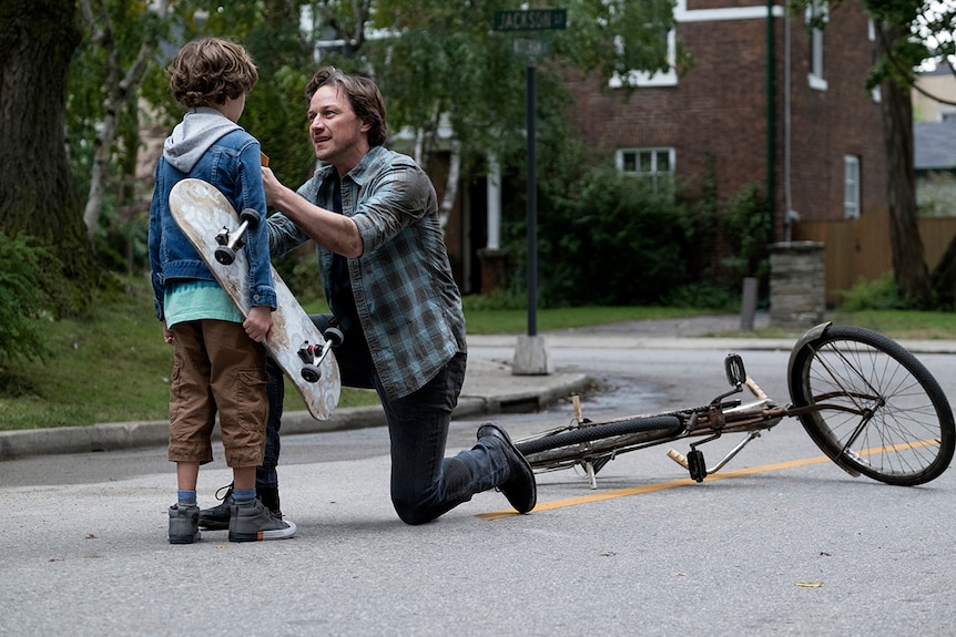 A man kneels on one knee near a fallen bike in a suburban street and holds onto the jacket of a young boy holding skateboard.