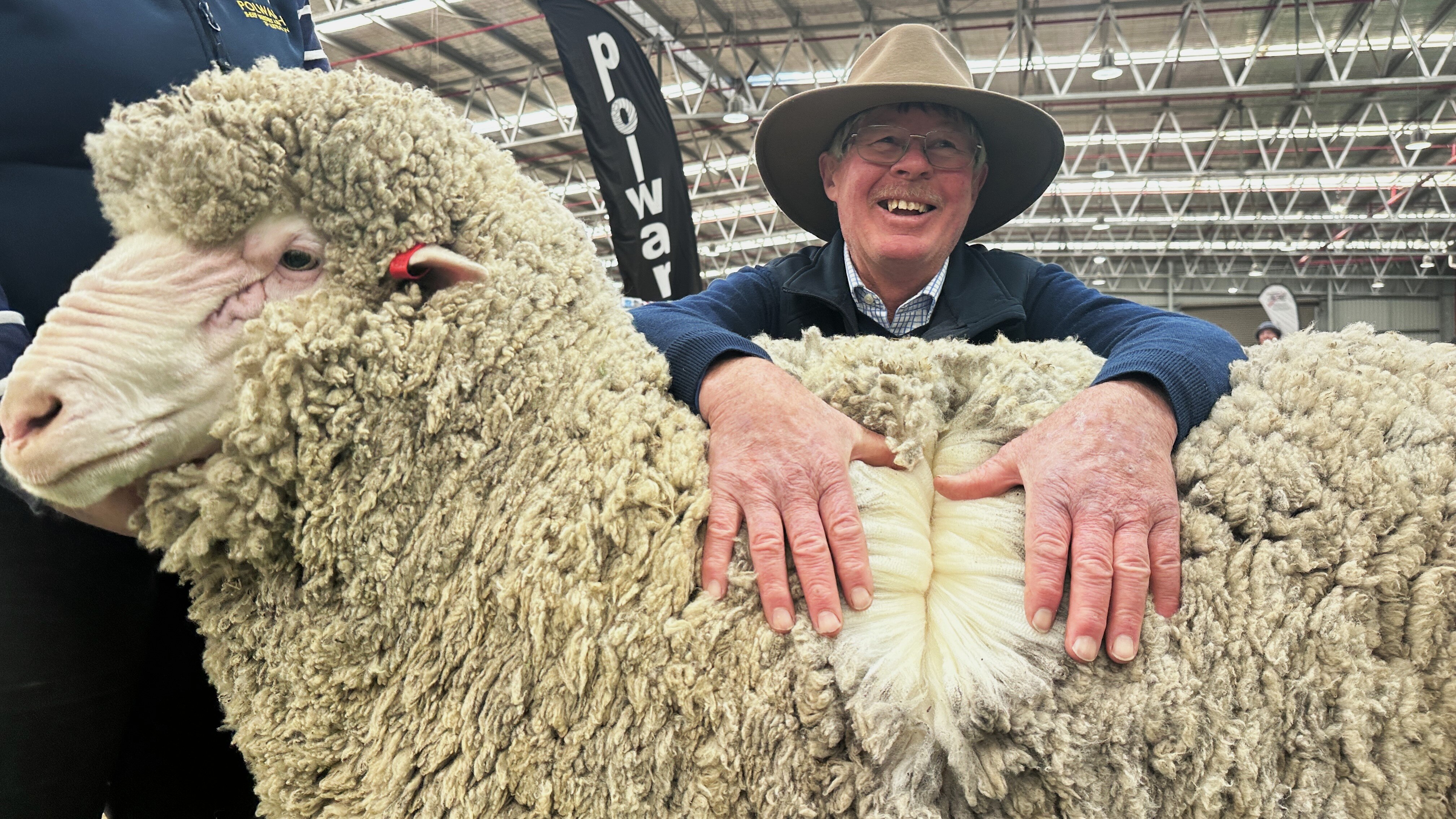 A man in an Akubra-style hat smiles and rests his hands on a sheep's fleece.