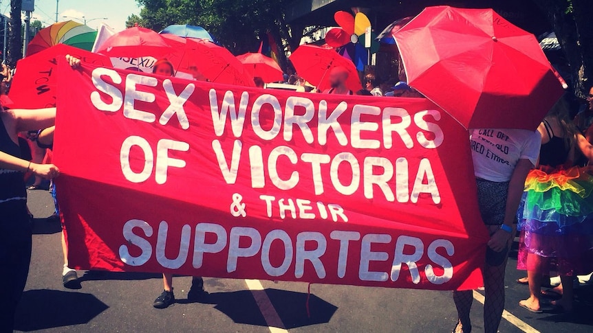 People march down a street in the sunlight holding a red sign reading 'SEX WORKERS OF VICTORIA & THEIR SUPPORTERS'.