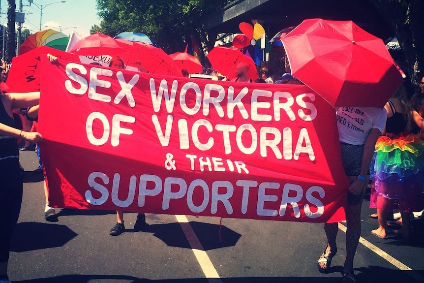People march down a street in the sunlight holding a red sign reading 'SEX WORKERS OF VICTORIA & THEIR SUPPORTERS'.