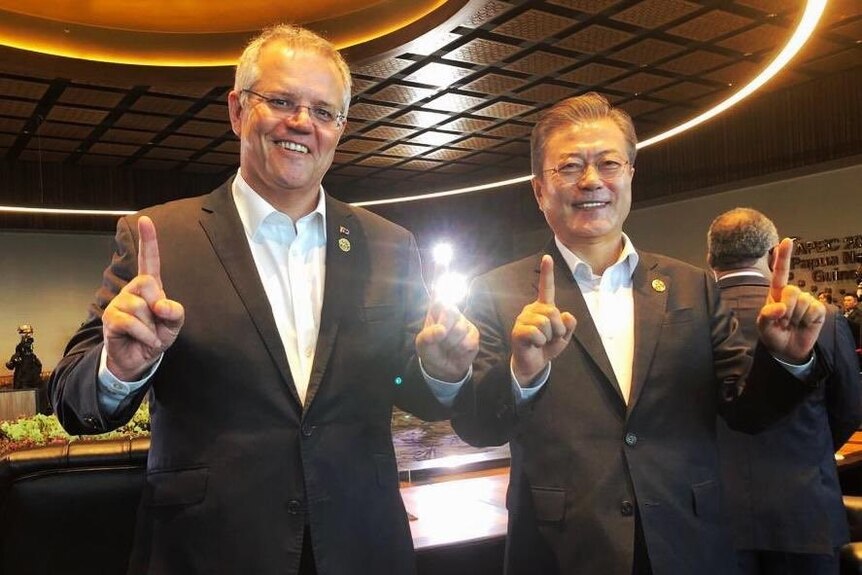 Two man in suits standing next to each other, holding up their index fingers