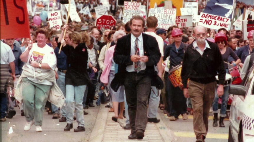 A man in a coat and tie walks in front of a huge crowd carrying placards and banners.