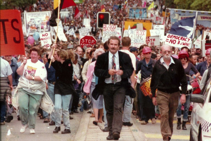 A man in a coat and tie walks in front of a huge crowd carrying placards and banners.