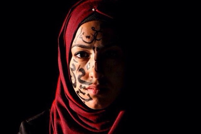 A woman wearing a red hijab with arabic writing on one side of her face in a dark room. 