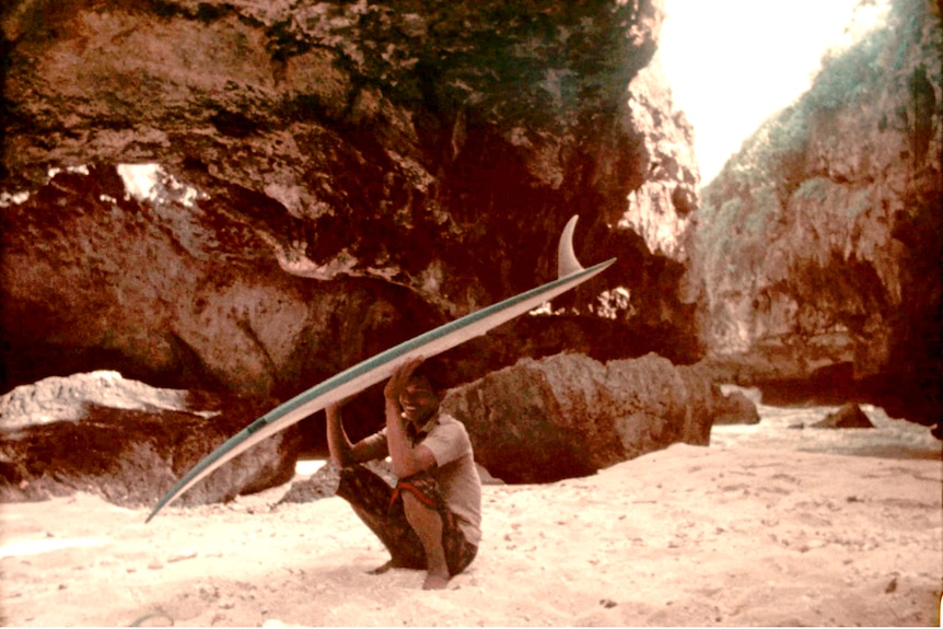 A man crouches in a cave in Uluwatu, holding a surfboard over his head.