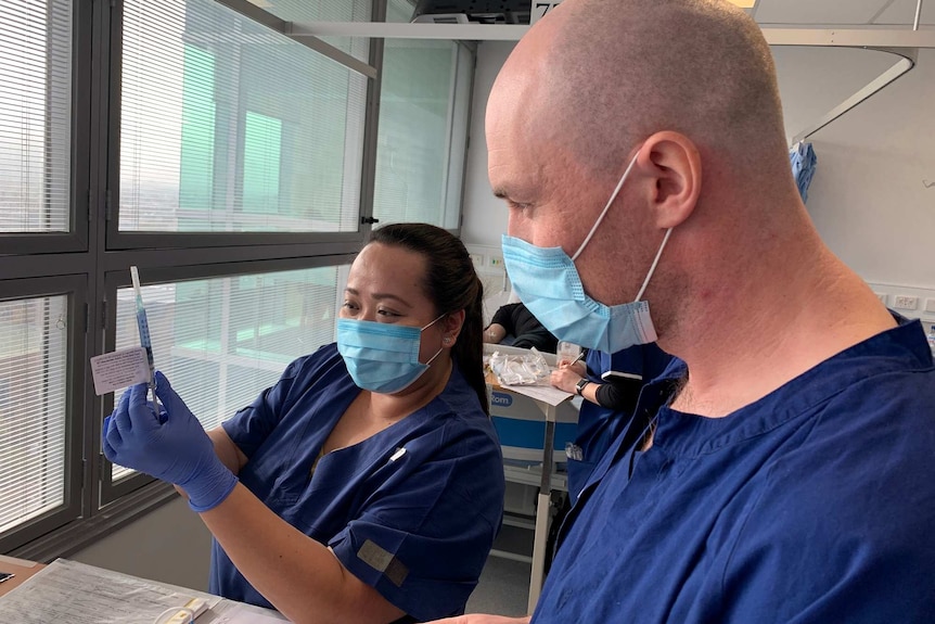 A woman and man in blue scrubs and masks prepare a syringe with a vaccine.