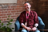 A man sits on a chair smiling while he is being interviewed