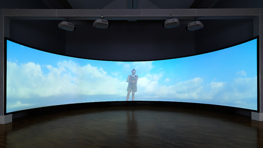 Gallery space showing curved video screen with figure of young man standing against a blue sky with clouds.