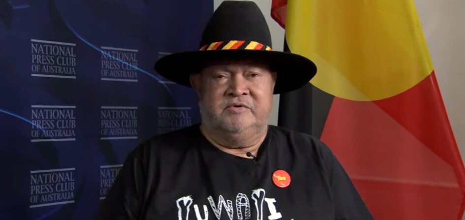 Senator Patrick Dodson sits in front of an Aboriginal flag, wearing a wide brim hat with a red, yellow and black stripped band. 