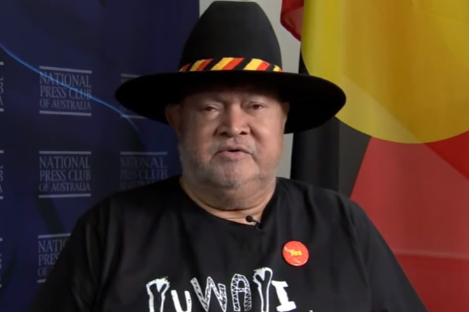 Senator Patrick Dodson sits in front of an Aboriginal flag, wearing a wide brim hat with a red, yellow and black stripped band. 