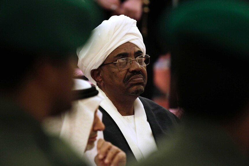 You see a close-up of Omar al-Bashir wearing white head robes as soft focus blurs two military guards on either side of his head