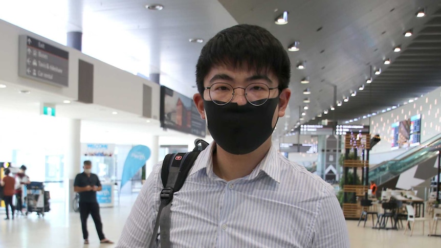 A man stands in an airport wearing a black face mask.
