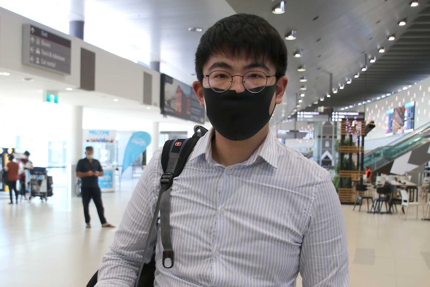 A man stands in an airport wearing a black face mask.