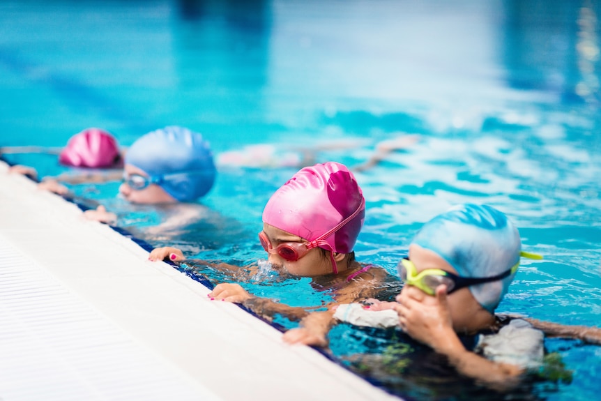 four children, each wearing blue and pink swimming caps, are in a swimming pool gripping the edge of the pool