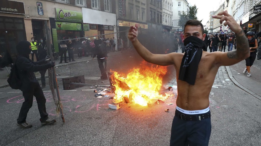 A shirtless man with a scarf around his mouth holds up his arms. There is fire in the background.