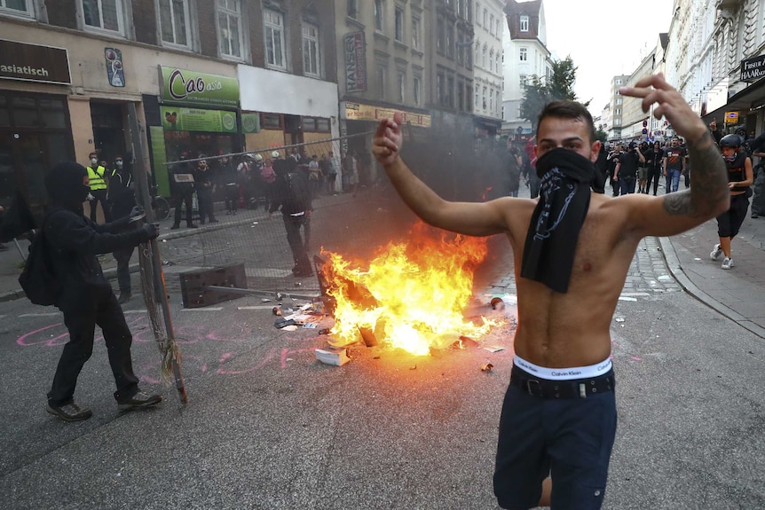 A shirtless man with a scarf around his mouth holds up his arms. There is fire in the background.