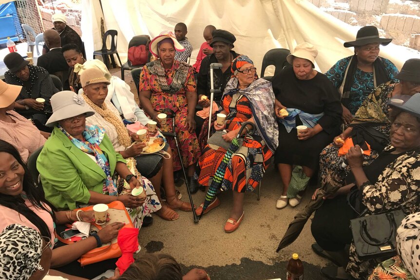 A group of African women sit in a circle in a tent.