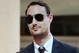 A young man in a suit and sunglasses holding an unseen woman's arm