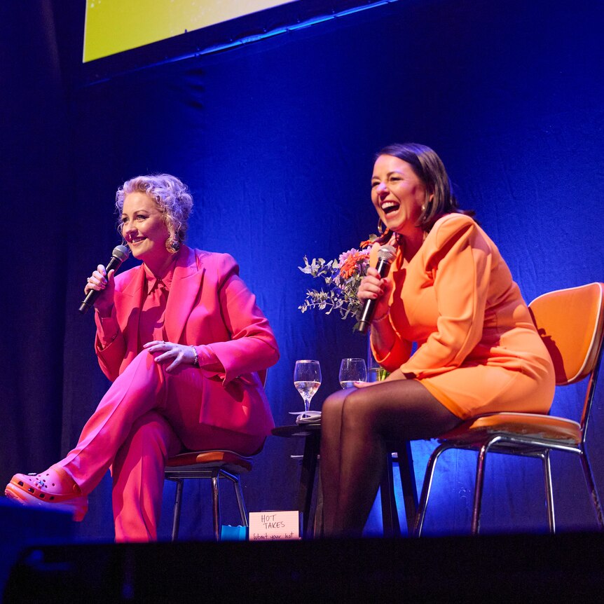 Zan and Myf sit on stage wearing pink and orange suits. They're laughing and holding microphones