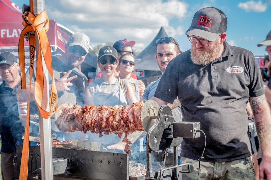 A man in glasses and a cap turns meat on a smoking pit in front of smiling onlookers at the 2019 Melbourne Meatstock event.