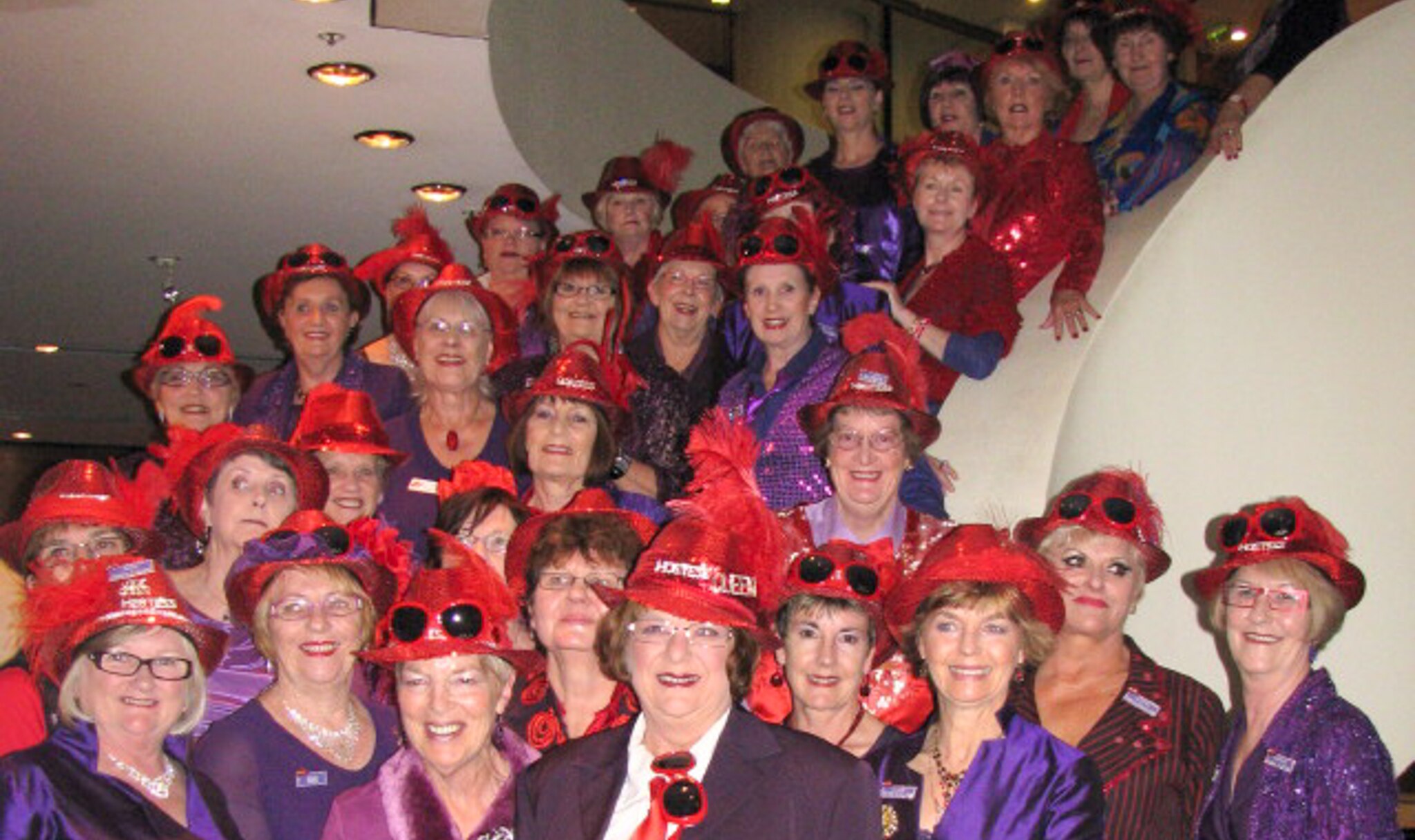 A large group of women stand on a twisted staircase wearing red hats and purple dresses.