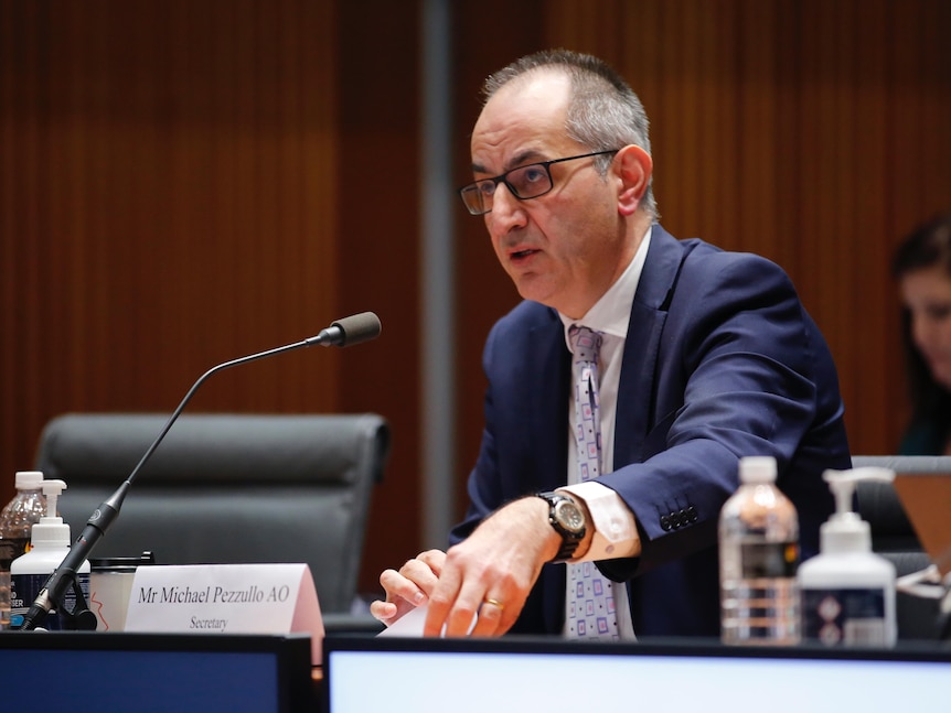 A man with short grey hair and glasses wearing a suit, sitting behind a microphone icking up a piece of paper as he speaks