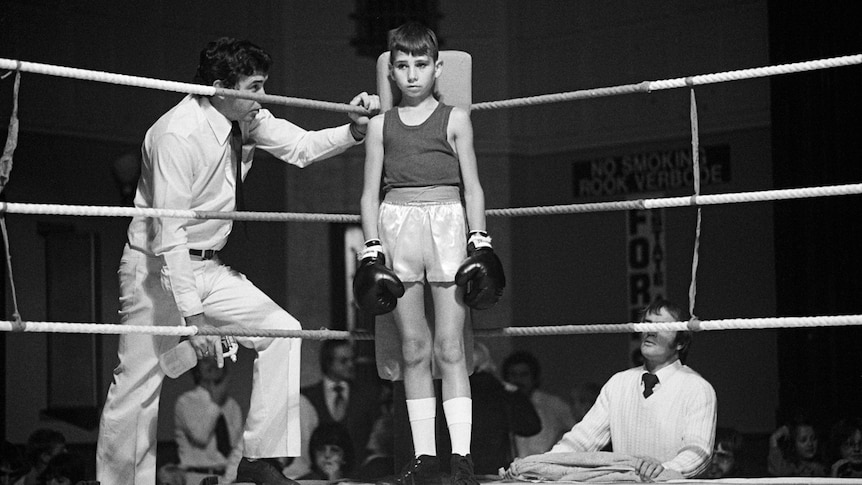 Small boy in boxing ring, facing camera, man on either side, crowd just visible behind them. Black and white photo.