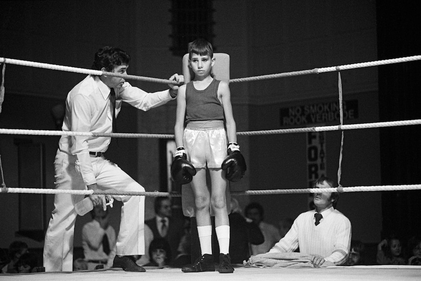 Small boy in boxing ring, facing camera, man on either side, crowd just visible behind them. Black and white photo.