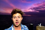 Young man with short brown hair and a button-up shirt with flowers on it, on a boat with purple sky background.