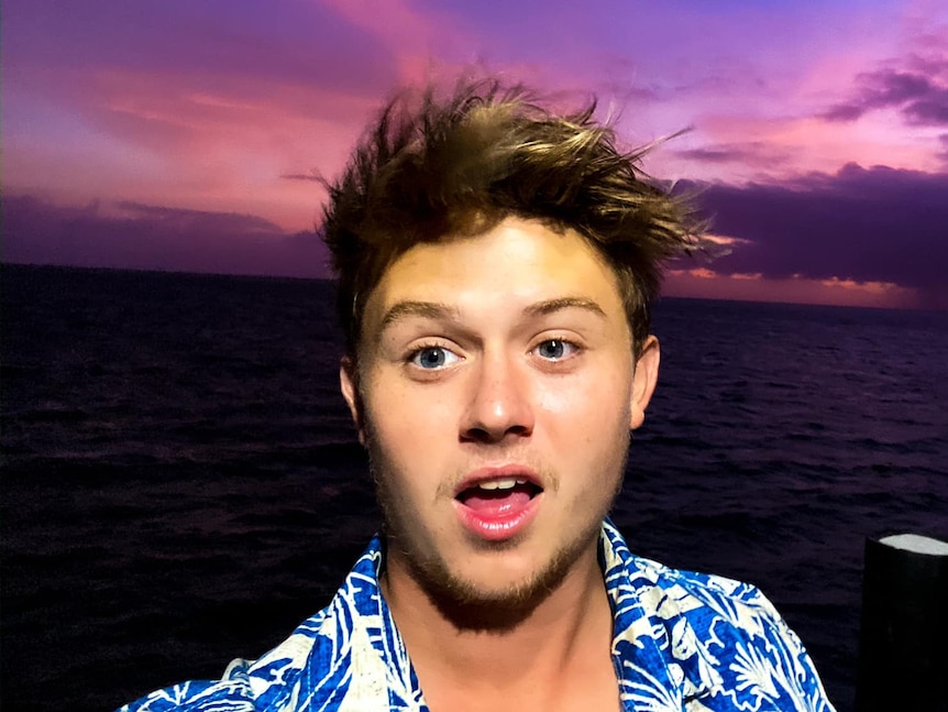 Young man with short brown hair and a button-up shirt with flowers on it, on a boat with purple sky background.