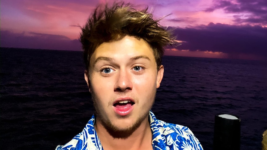 Young man with short brown hair and a button-up shirt with flowers on it, on a boat with purple sky background.